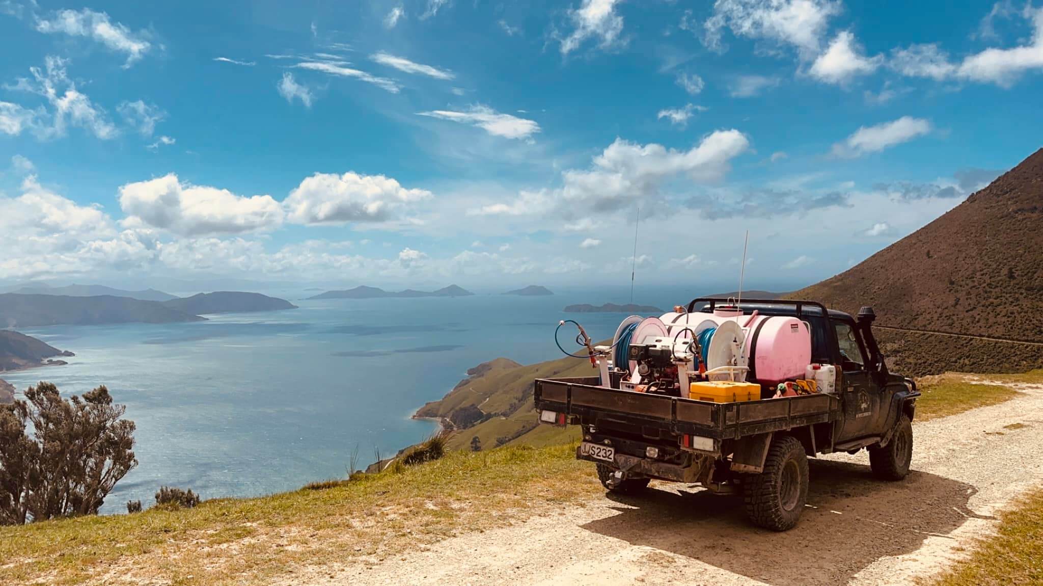 A truck loaded with equipment is parked on a dirt road overlooking a coastal landscape with hills and a body of water under a blue sky.
