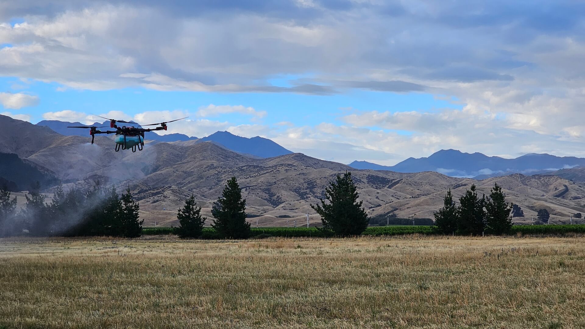 A drone spraying a field with mountains in Marlborough and trees in the background under a cloudy sky.