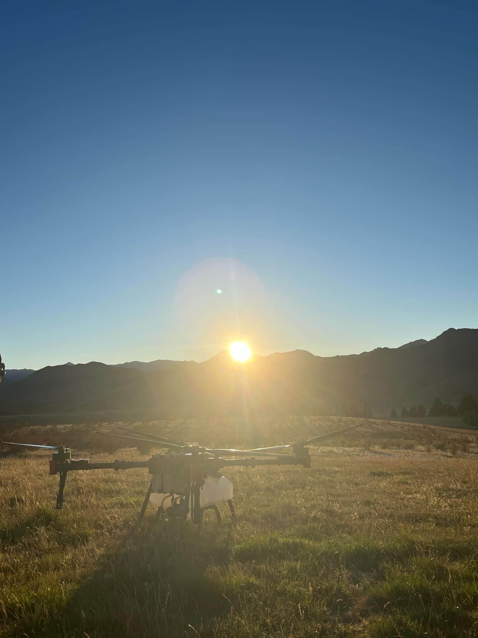 A drone rests on grassy terrain at sunset, with mountains silhouetted against a clear sky in the background.