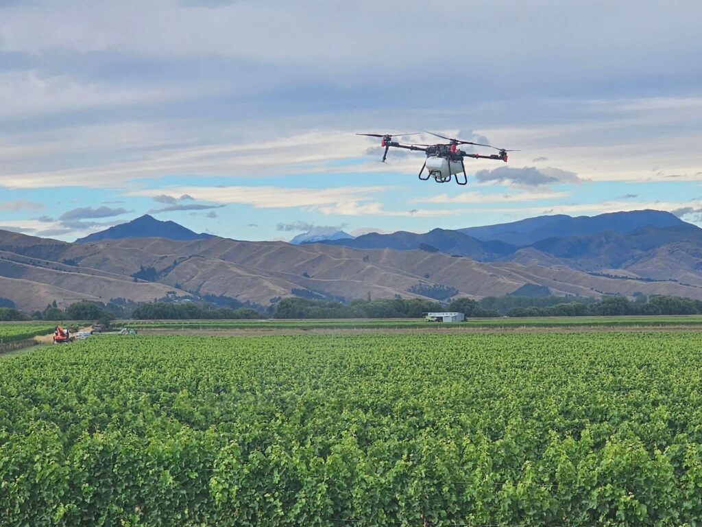 A drone flying over a lush green vineyard set against a backdrop of rolling hills and a partly cloudy sky.