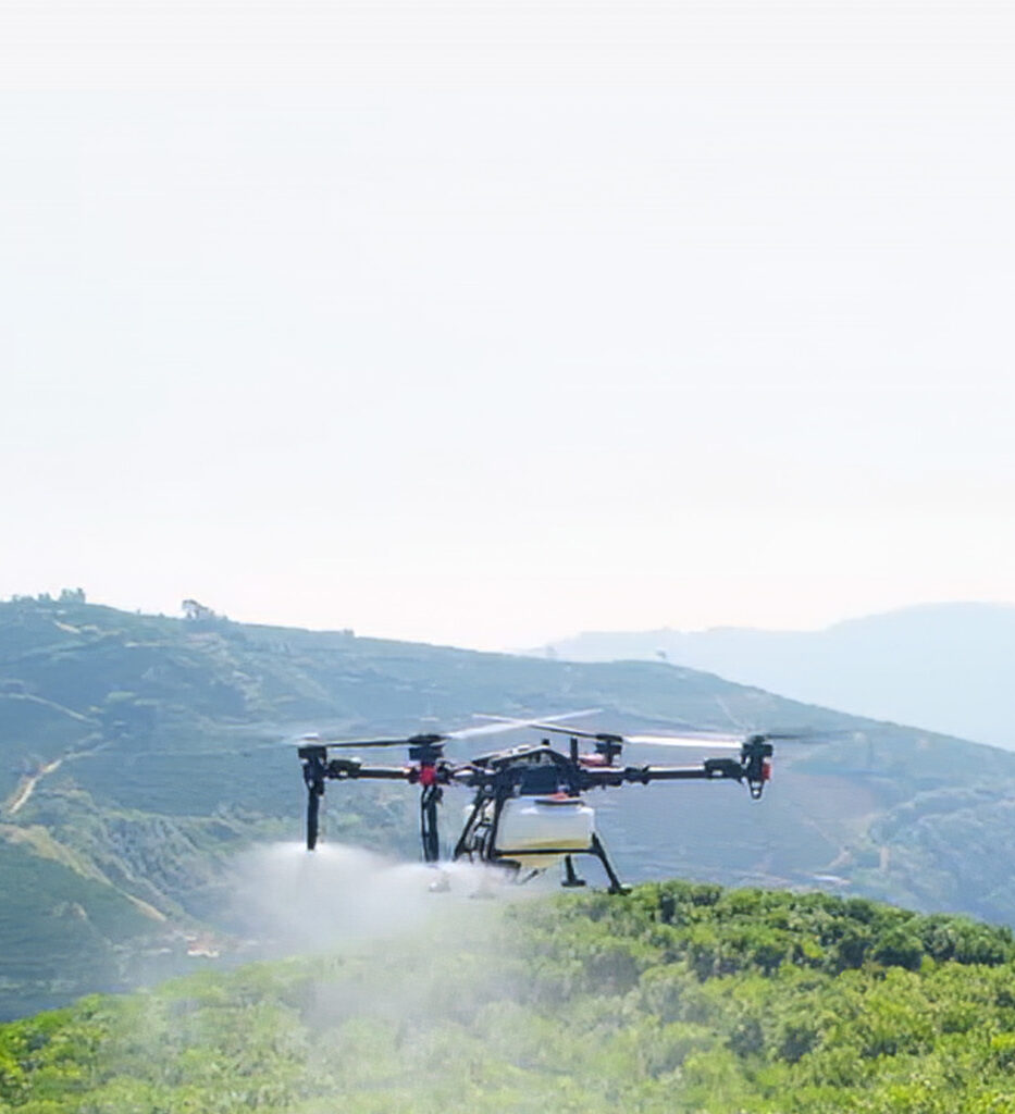 A drone sprays crops in a green field with hills and mountains visible in the background under a clear sky.